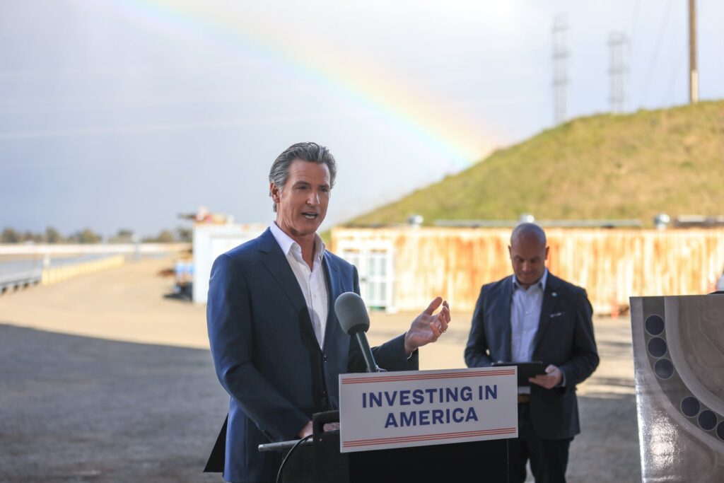 California Governor Gavin Newsom speaking outdoors at a lectern marked “Investing in America”