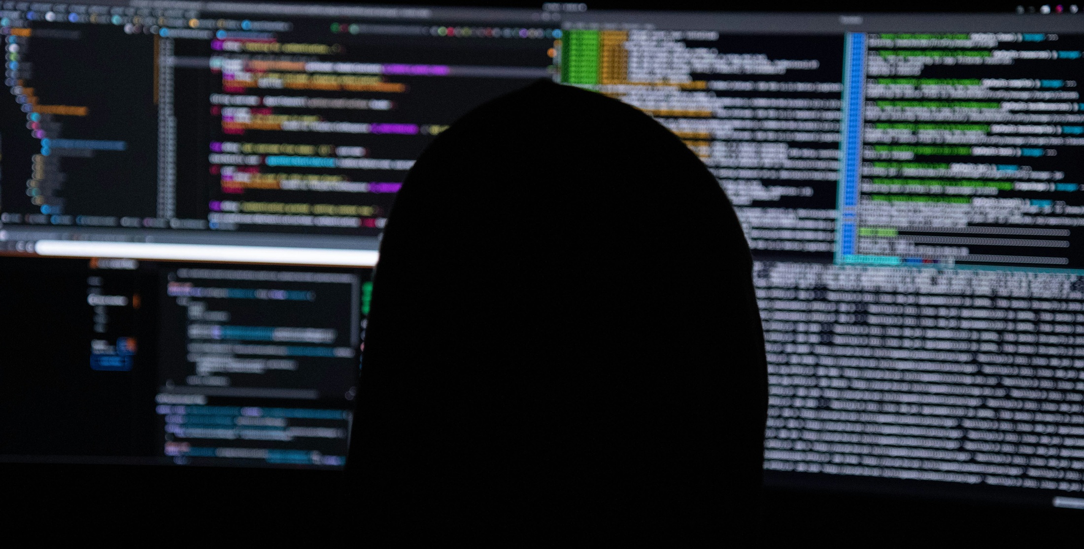Hooded figure silhouetted against monitors full of code