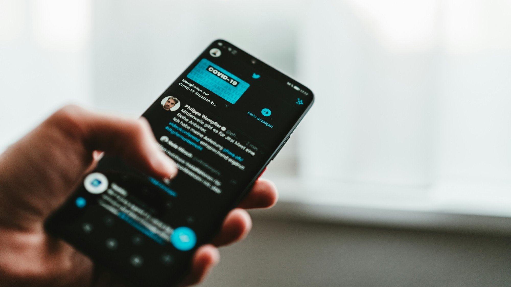 A hand holding a phone showing the Twitter app in dark mode, with a feed of tweets on screen.