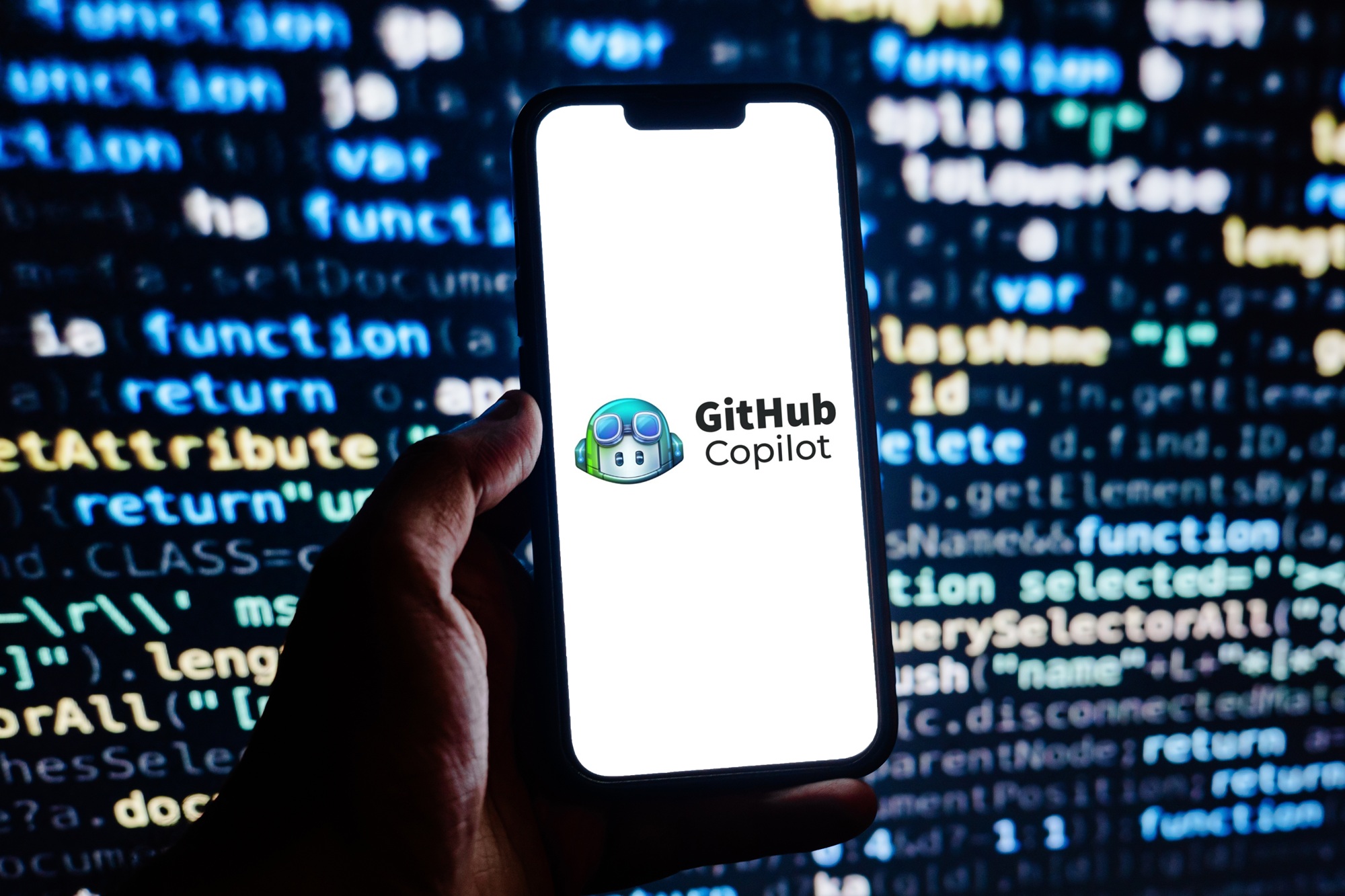 A phone showing the GitHub Copilot logo, held in front of a screen of blurred code