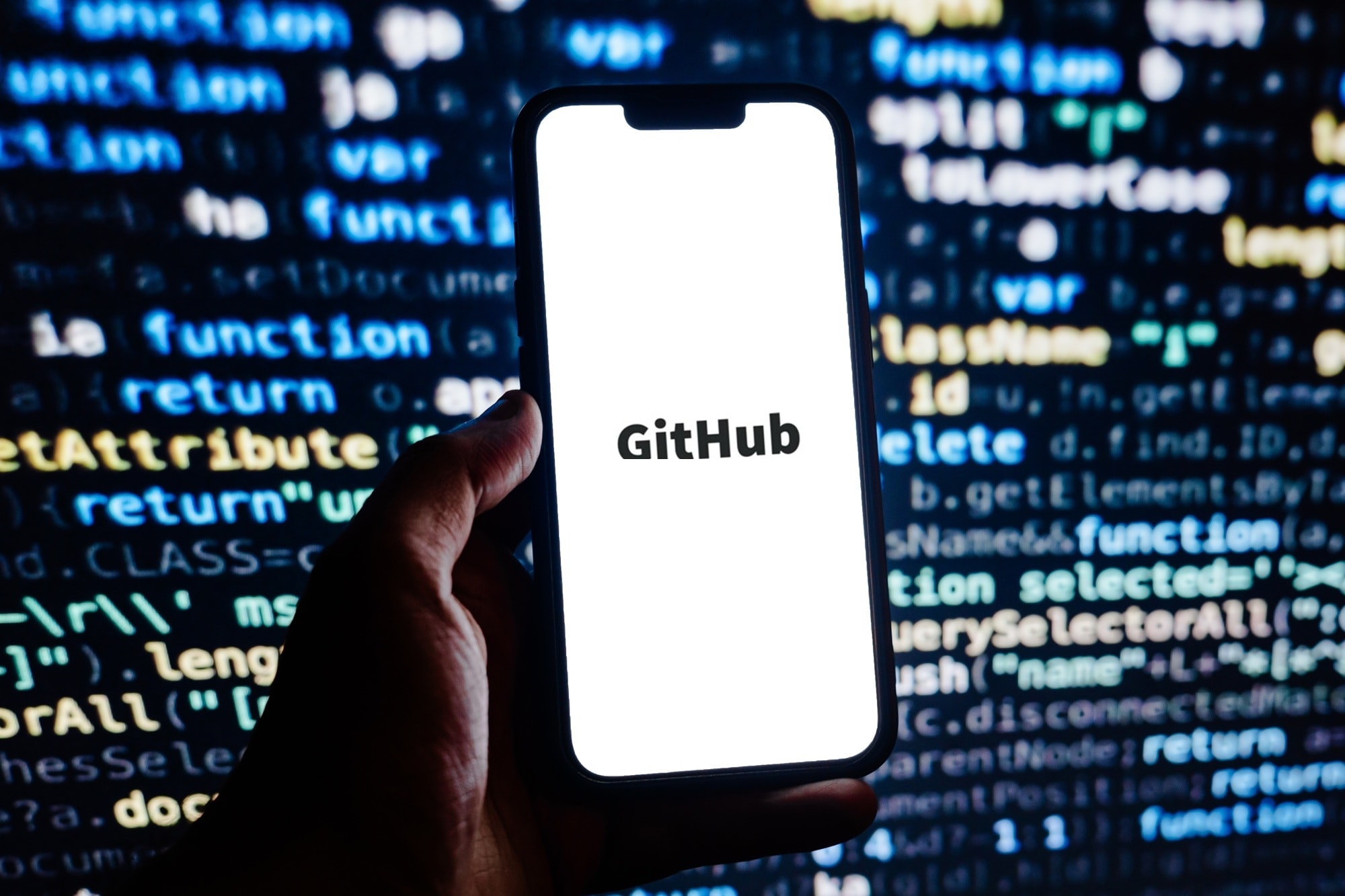 A phone showing the word GitHub, held in front of a screen of blurred code