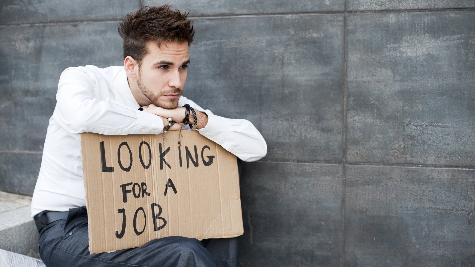 A man in a white shirt sitting on a step, leaning on a cardboard sign that reads Looking for a job