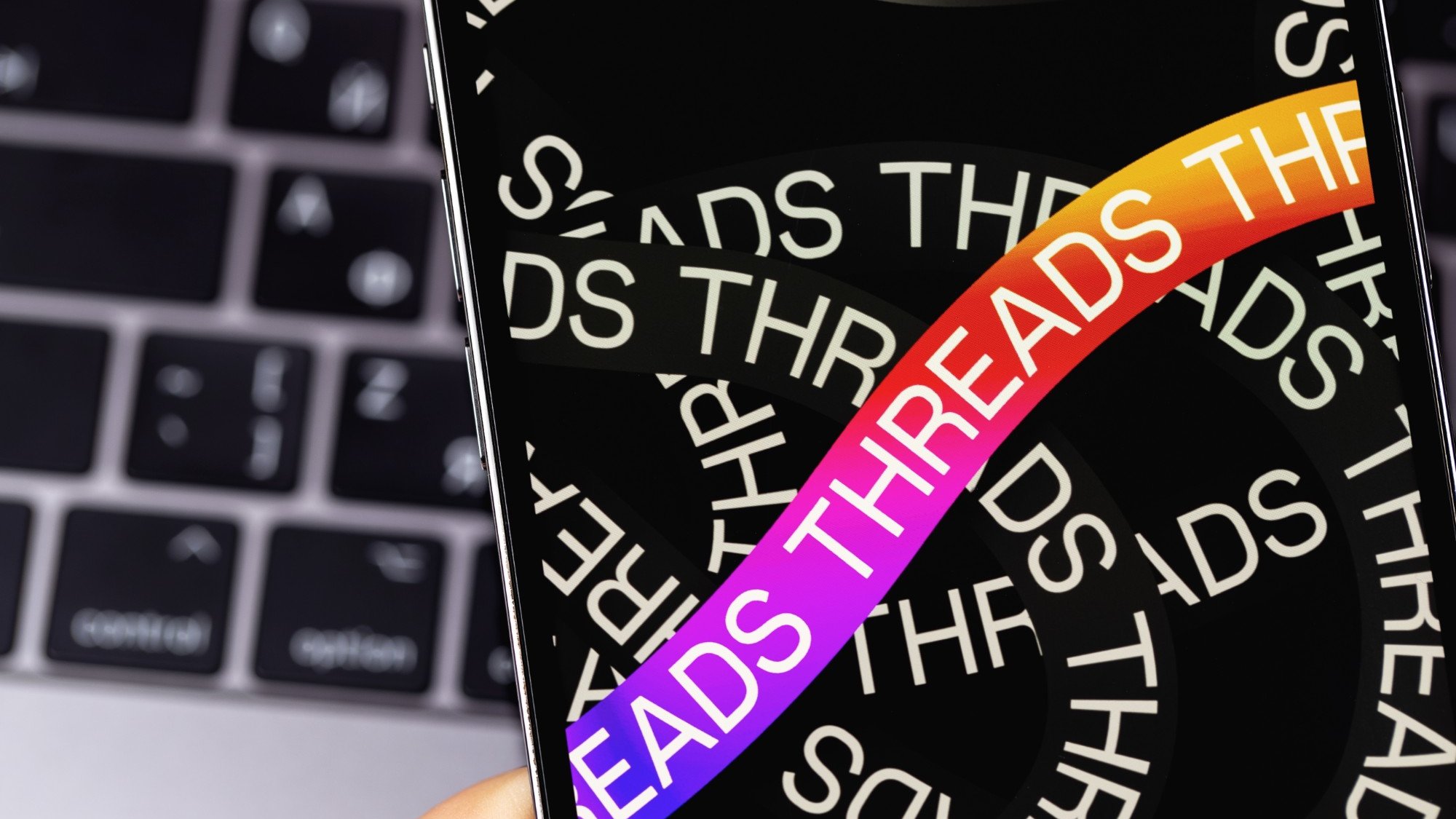 A phone showing the Threads app's launch screen, held in front of a laptop keyboard