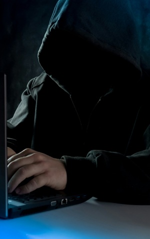 A hooded figure at a laptop in the dark, face hidden, hands on the keyboard